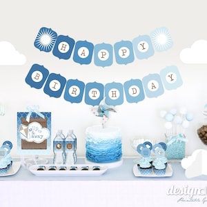 Op de afbeelding: Een blauwe en bruine gedecoreerde tafel voor een verjaardagsfeest met een taart, cupcakes en andere lekkernijen. De banner luidt "Happy Birthday".