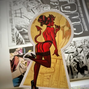 May include: A sticker featuring a red devil woman in a keyhole design, applying lipstick. The illustration includes a tail, horns, and black lingerie. The background shows comic book pages. The sticker has the artist's name, ARTISTABE, at the bottom.