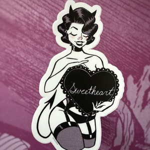 May include: A black and white sticker featuring a pin-up style illustration of a woman with devil horns holding a heart-shaped sign. The sign reads "Sweetheart" in cursive. The sticker has a white border and is set against a purple background.