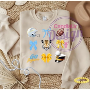 May include: A beige sweatshirt with various graphics, including a football helmet, bows, a jaguar face, a football, a lightbulb, and a military hat. The text "Go Jaguars" is also visible. The design is likely for a sports team or school.