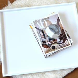 May include: A large square white tray with raised edges. Resting on the tray is a stack of Architectural Digest magazines featuring the text "AT HOME WITH JULIANNE MOORE THE MOVIE STAR NEXT DOOR" and a large geometric gray and white stone object.