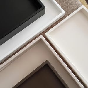 May include: A collection of three rectangular trays arranged in an overlapping pattern. One tray features a matte black interior with a white border, while the other two trays are solid white and off-white with raised edges and smooth surfaces.