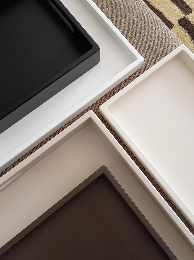 May include: A collection of three rectangular trays arranged in an overlapping pattern. One tray features a matte black interior with a white border, while the other two trays are solid white and off-white with raised edges and smooth surfaces.