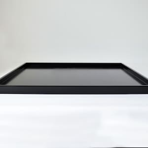 Może przedstawiać: Czarna, kwadratowa taca do serwowania o niskim profilu. Taca ma prosty, nowoczesny design z płaską powierzchnią i podniesionymi krawędziami. Taca wykonana jest z solidnego, ciemnego materiału i nadaje się do serwowania jedzenia lub eksponowania przedmiot&oacute;w.