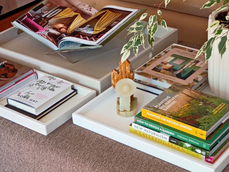 May include: Two white rectangular serving trays holding various books and decorative items. One tray contains The Book of Delights by Ross Gay. The other tray holds a stack of gardening books, a small stone cactus sculpture, and a white plant pot.