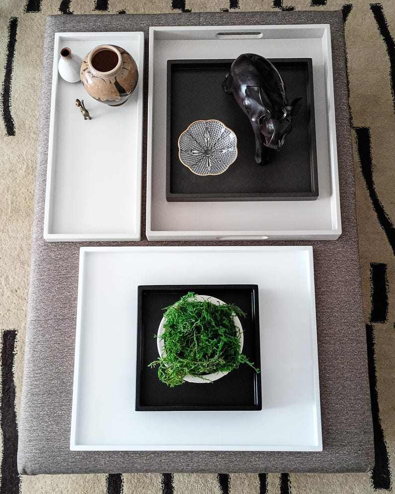 May include: A collection of rectangular and square trays in white, light gray, and black. The trays hold a ceramic vase, a small gold figurine, a black animal sculpture, a patterned bowl, and a white bowl filled with bright green moss.