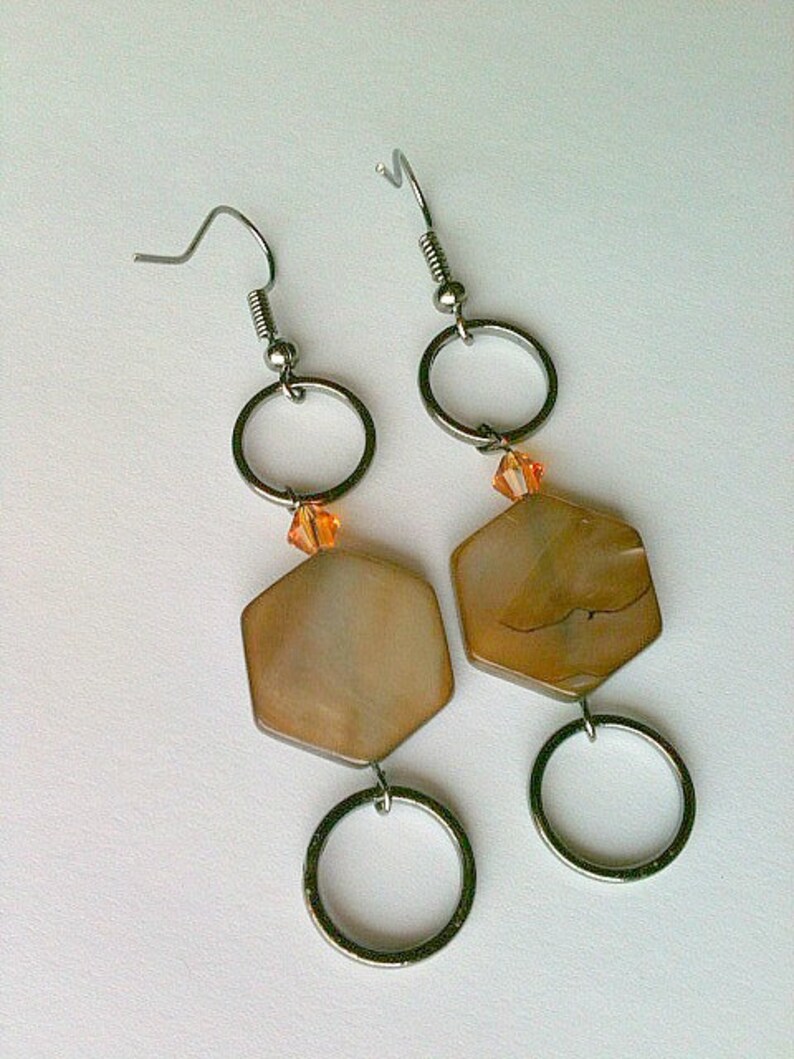 May include: A pair of silver earrings with a hexagonal shell-like pendant and a small orange crystal bead. The earrings have a silver hoop and a silver hook.