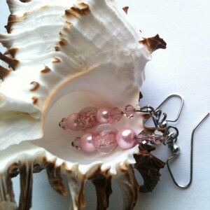 May include: Pink and clear glass bead earrings with silver hooks. The earrings are displayed in a seashell.