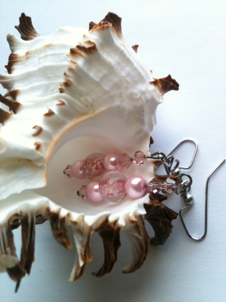 May include: Pink and clear glass bead earrings with silver hooks. The earrings are displayed in a seashell.