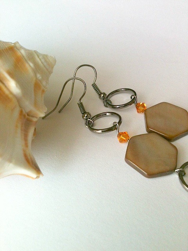 May include: A pair of earrings with a hexagonal shaped pendant made of brown mother of pearl. The earrings have a silver hook and a small orange crystal bead.