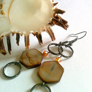 May include: A pair of earrings with hexagonal shell-shaped pendants, brown and white in color, with a dark gray metal hoop and a small amber-colored bead on each earring.
