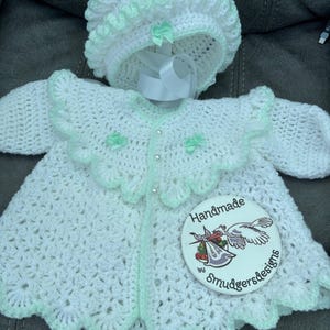 Puede incluir: Conjunto de bebé de ganchillo blanco hecho a mano con gorro a juego. El conjunto presenta un borde festoneado en verde menta suave, acentuado con pequeños lazos. Incluye una chaqueta con botones de perlas y una etiqueta redonda que dice "Handmade by Smudgersdesigns".
