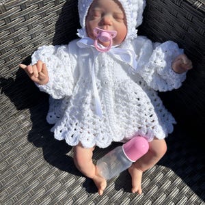 May include: A baby doll dressed in a white crocheted outfit, featuring a bonnet and dress, with a pink dummy and bottle. The doll is lying on a woven surface, with shadows from the sun.