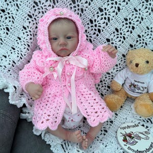 Könnte beinhalten: Eine lebensechte Babypuppe in einem handgefertigten, rosa gehäkelten Kapuzenjäckchen mit Perlenakzenten und passender Kapuze. Ein kleiner Teddybär in einem weißen T-Shirt sitzt daneben. Die Puppe liegt auf einer weißen Häkeldecke.