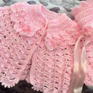 Könnte beinhalten: Ein rosa gehäkeltes Baby-Jäckchen mit Rüschenkragen und passender Mütze. Das Jäckchen hat einen Knopfverschluss und die Mütze ein Satinband.