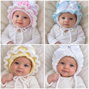 May include: A collection of four crocheted baby bonnets in pink, light blue, yellow, and white. Each bonnet features a ruffled edge, a satin ribbon bow at the top center with a small crystal accent, and matching crocheted ties under the chin.