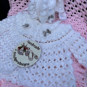 Peut inclure: Ensemble de robe et bonnet pour bébé en crochet blanc avec une couverture en crochet rose. La robe a un bord festonné et le bonnet a un volant. La couverture a un motif de point ouvert simple. L'ensemble est fait à la main par Smulders Designs.