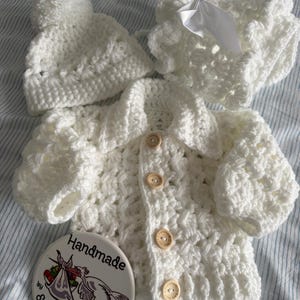 May include: A white crocheted set consisting of a long-sleeved cardigan with four light brown wooden buttons, a beanie with a pom-pom, and a ruffled bonnet with a white satin bow. A circular tag features a stork illustration and the text Handmade by Smudgersdesigns.