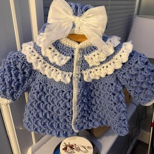 May include: A light blue crocheted cardigan with a white ruffled collar and white trim on the sleeves. The garment features three clear buttons down the front and is topped with a large white satin bow. A circular tag displays the text Smudgers.