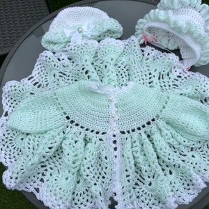 May include: A crocheted baby outfit in white and light green. The outfit includes a cardigan, a bonnet, and a matching hat.