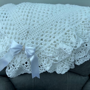 May include: A white, crocheted blanket with a scalloped edge and a satin bow. The blanket has a delicate, open weave pattern. The bow is adorned with a small, sparkly embellishment. The blanket is folded over a dark surface.