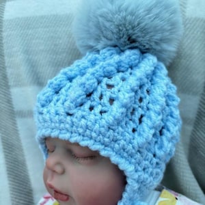 Peut inclure: Un bonnet de bébé en crochet bleu clair avec un gros pompon gris duveteux. Le bonnet présente un motif texturé et un bord. Le bébé porte une chemise à motifs floraux.