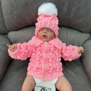 May include: A baby doll dressed in a handmade pink crochet hat with a white pom-pom and a matching pink crochet jacket. The jacket has a collar and is buttoned up. The doll is lying on a grey surface.