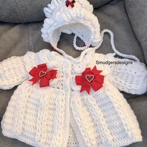 Op de afbeelding: Een witte gehaakte babyoutfit, inclusief een muts en een vest. De muts heeft een rode strik en het vest heeft twee rode strikken met hartvormige details. De outfit wordt gepresenteerd op een grijze ondergrond.