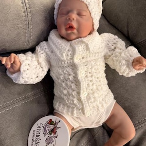 May include: A baby doll dressed in a white crocheted hat, cardigan, and onesie. The cardigan features a collar and button fastening. A round white object with the text "Smudders Designs" and a bird graphic is visible.