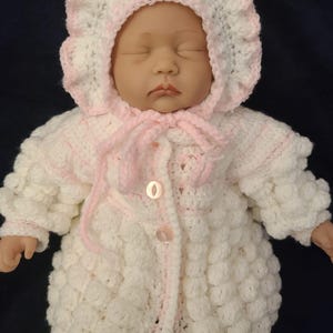 May include: A baby doll dressed in a hand-crocheted outfit. The outfit comprises a bonnet and a coat, both in white with pink detailing. The coat features a textured, bobble-like pattern and is fastened with buttons. The bonnet has a frilled edge and ties under the chin.