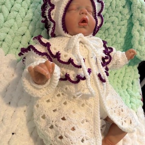 Puede incluir: Un conjunto de bebé de ganchillo blanco con ribetes morados, que incluye un gorro y un abrigo. El gorro tiene un lazo blanco. El bebé está acostado sobre una manta blanca y verde.