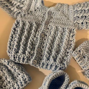 Może przedstawiać: Jasnoniebieski zestaw dla dziecka wykonany na szydełku, w tym rozpinany kardigan, dwie czapki i para bucików. Zestaw wykonany jest z miękkiej przędzy o wzorze ściegu. Ubranka dla dzieci są prezentowane na jasnej powierzchni.