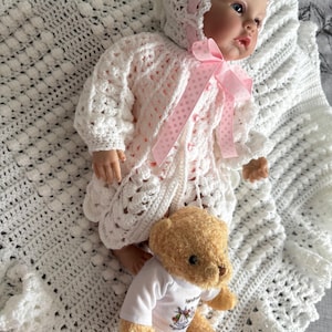 May include: A baby doll wearing a white crochet outfit with a matching bonnet and a pink ribbon. A small teddy bear in a white t-shirt is lying next to the doll on a white crocheted blanket.