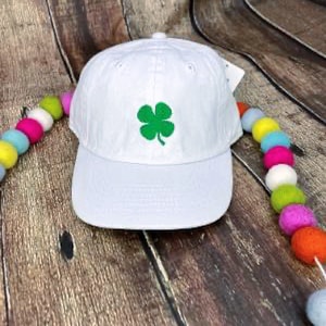 May include: A white baseball cap with a green four-leaf clover embroidered on the front. The cap has a curved brim and a fabric strap closure. The hat is displayed on a wooden surface.