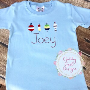 May include: Light blue short-sleeve t-shirt with embroidered buoys in red, yellow, green, and white. The name "Joey" is embroidered in red script. A floral logo with the text "Gabby Girl Designs" is in the lower right corner.