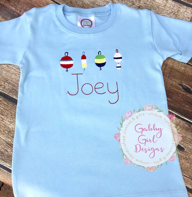 May include: Light blue short-sleeve t-shirt with embroidered buoys in red, yellow, green, and white. The name "Joey" is embroidered in red script. A floral logo with the text "Gabby Girl Designs" is in the lower right corner.