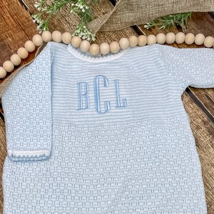 May include: A light blue baby romper with long sleeves and a textured weave. The romper features a striped upper section and a lower section with a woven pattern. The initials "BCL" are embroidered in light blue on the chest.