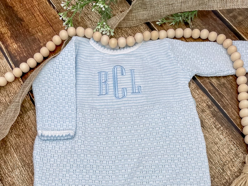 May include: A light blue baby romper with long sleeves and a textured weave. The romper features a striped upper section and a lower section with a woven pattern. The initials "BCL" are embroidered in light blue on the chest.