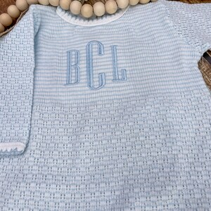 May include: Light blue baby outfit with the embroidered initials "BCL". The outfit features a striped pattern on the body and a textured knit on the sleeves. The neckline and sleeve cuffs are trimmed with white.