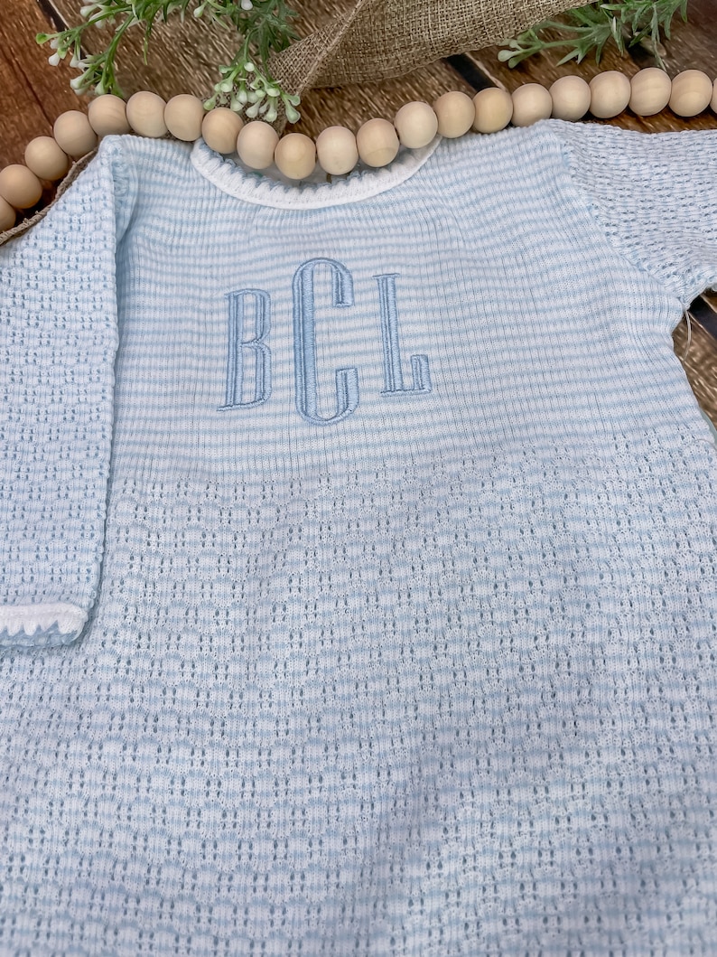 May include: Light blue baby outfit with the embroidered initials "BCL". The outfit features a striped pattern on the body and a textured knit on the sleeves. The neckline and sleeve cuffs are trimmed with white.