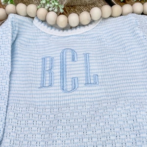 May include: A light blue and white striped baby sweater with long sleeves. The sweater features an embroidered monogram in light blue thread. The knitwear has a delicate openwork pattern on the sleeves and a scalloped neckline.