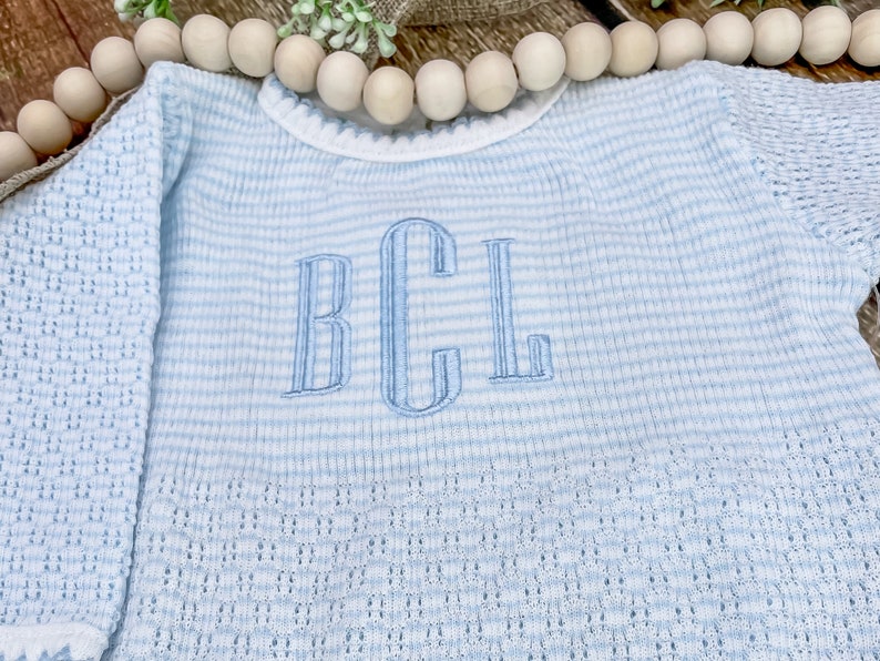 May include: A light blue and white striped baby sweater with long sleeves. The sweater features an embroidered monogram in light blue thread. The knitwear has a delicate openwork pattern on the sleeves and a scalloped neckline.