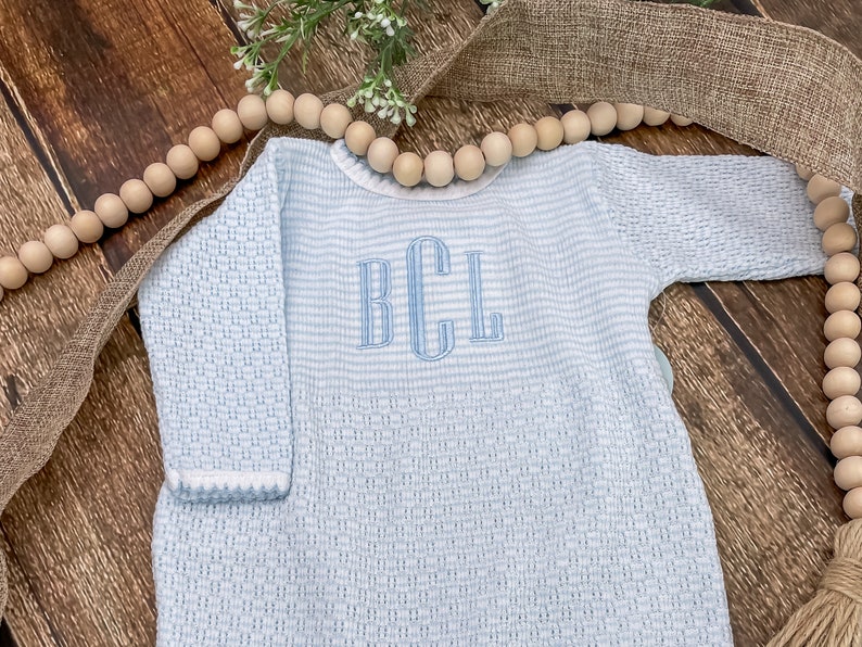 May include: A light blue baby romper with a textured weave and horizontal stripes. The romper features a white trim on the collar and cuffs, and a monogram in light blue thread. The background includes wooden beads and a burlap ribbon.