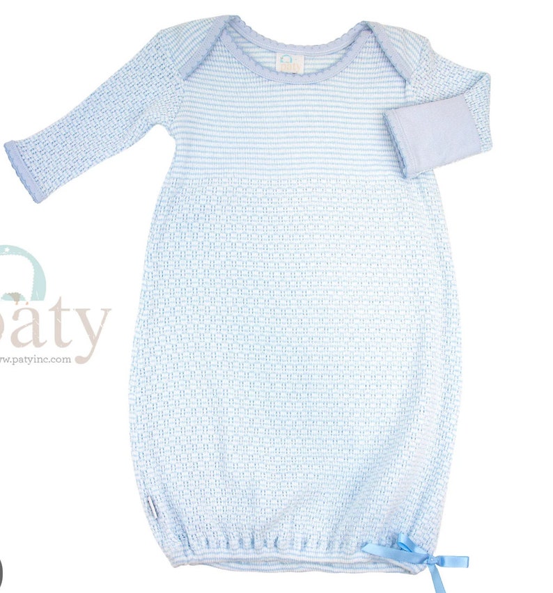 May include: A light blue baby gown with long sleeves and a woven pattern. The gown features a light blue trim around the neckline and cuffs, and a small bow at the bottom. The gown is made of a soft, knitted material.