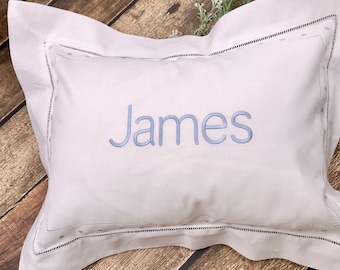 May include: White pillowcase with the name "James" embroidered in light blue. The pillowcase has a decorative border with small, evenly spaced dots and a stitched edge. The pillow is on a wooden surface.