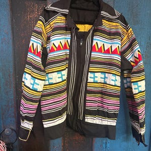 May include: A black zip-up jacket featuring a vibrant, multi-colored geometric pattern. The design includes horizontal bands of yellow, pink, and white zig-zag stitching, along with sections of red, yellow, and white triangles and blue squares.