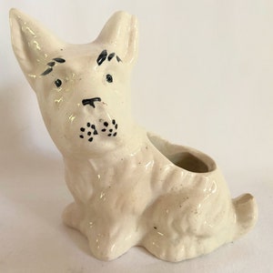Può includere: Vaso da fiori in ceramica bianca a forma di cane con dettagli dipinti in nero. Il cane è seduto con la testa inclinata e ha una piccola apertura nella schiena per piantare.