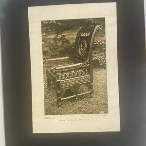 Op de afbeelding: Een zwart-witfoto van een houten fauteuil met ingewikkelde houtsnijwerk. De stoel staat in een tuin en heeft een plaquette op de rug met de tekst "The Children of the Poet". De onderschrift onder de afbeelding luidt "Longfellow's Armchair."