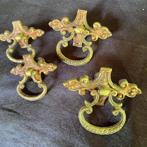 Puede incluir: Cuatro tiradores de cajón de latón antiguo con diseños ornamentados. Cada tirador tiene una manija en forma de anillo y una placa posterior decorativa en forma de cruz.