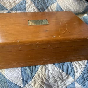 May include: A rectangular wooden box with a brass nameplate on the lid. The box is a warm, medium brown color and shows signs of wear. It sits on a blue and white patterned quilt.